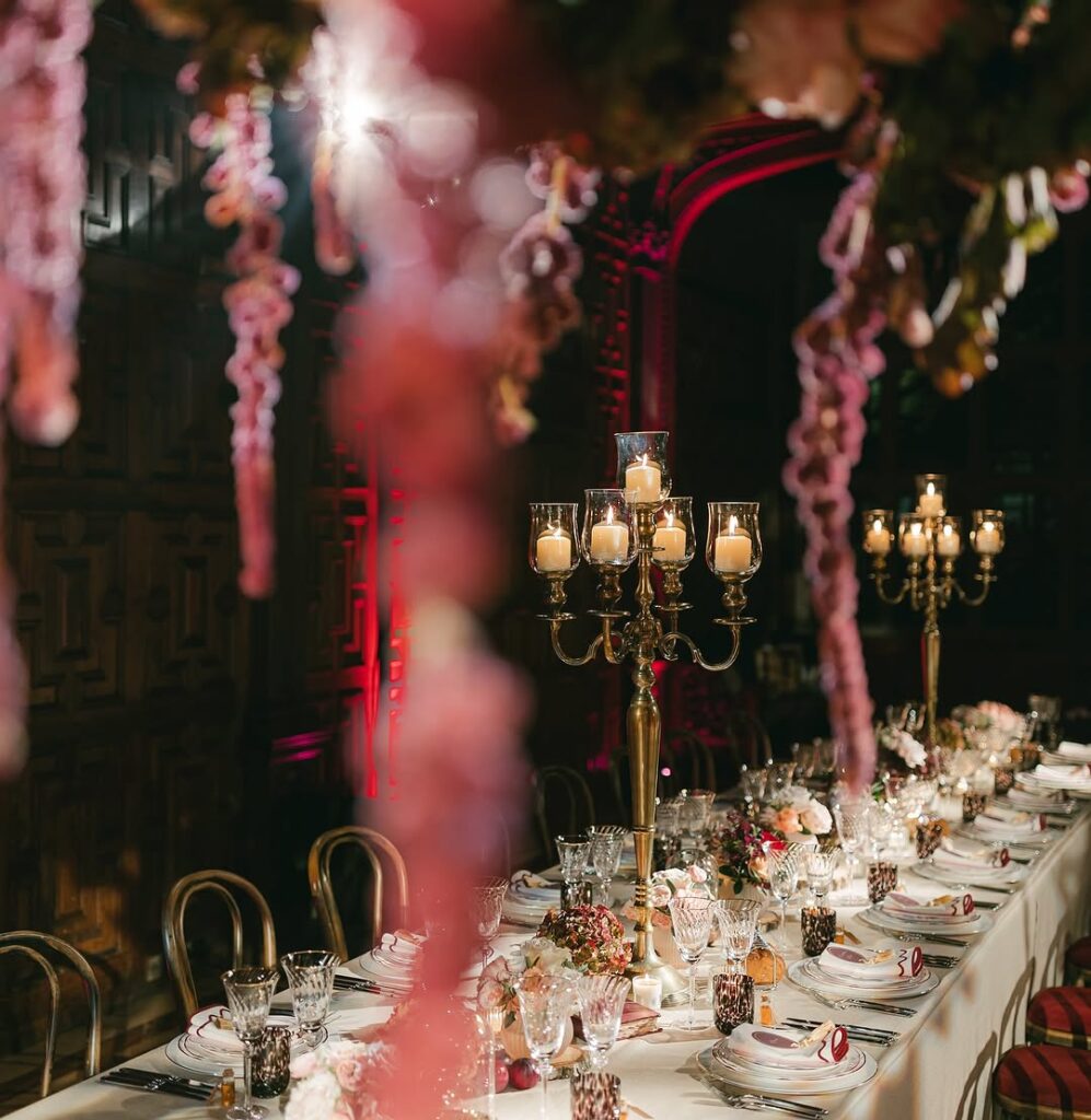 A luxurious dinner table setup at Two Temple Place with tall gold candelabras, warm candlelight, and floral arrangements cascading from above in soft pink and deep red hues.