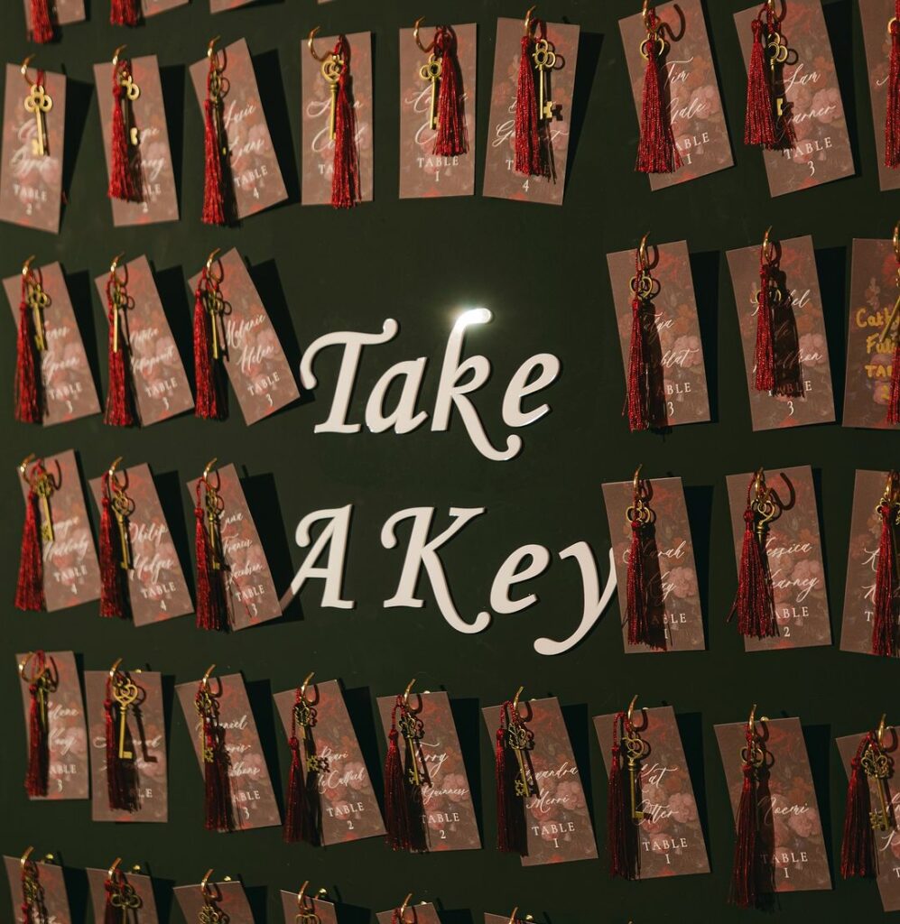 A unique seating chart display featuring gold keys with red tassels hanging on name cards, set against a dark background with the text “Take A Key.”