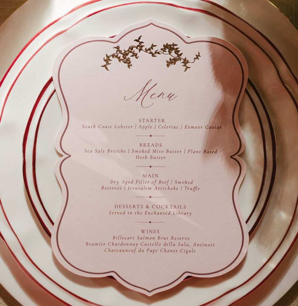 A detailed close-up of an elegant menu placed on layered plates with red accents. The menu lists the gourmet courses for the evening in refined typography with gold embellishments.