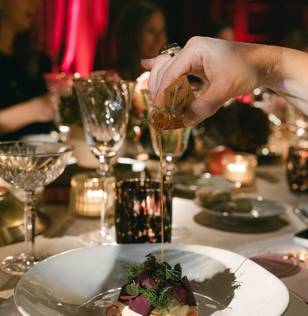 A guest pouring a small bottle of infused olive oil over a beautifully presented dish, surrounded by elegant glassware and soft candlelight.