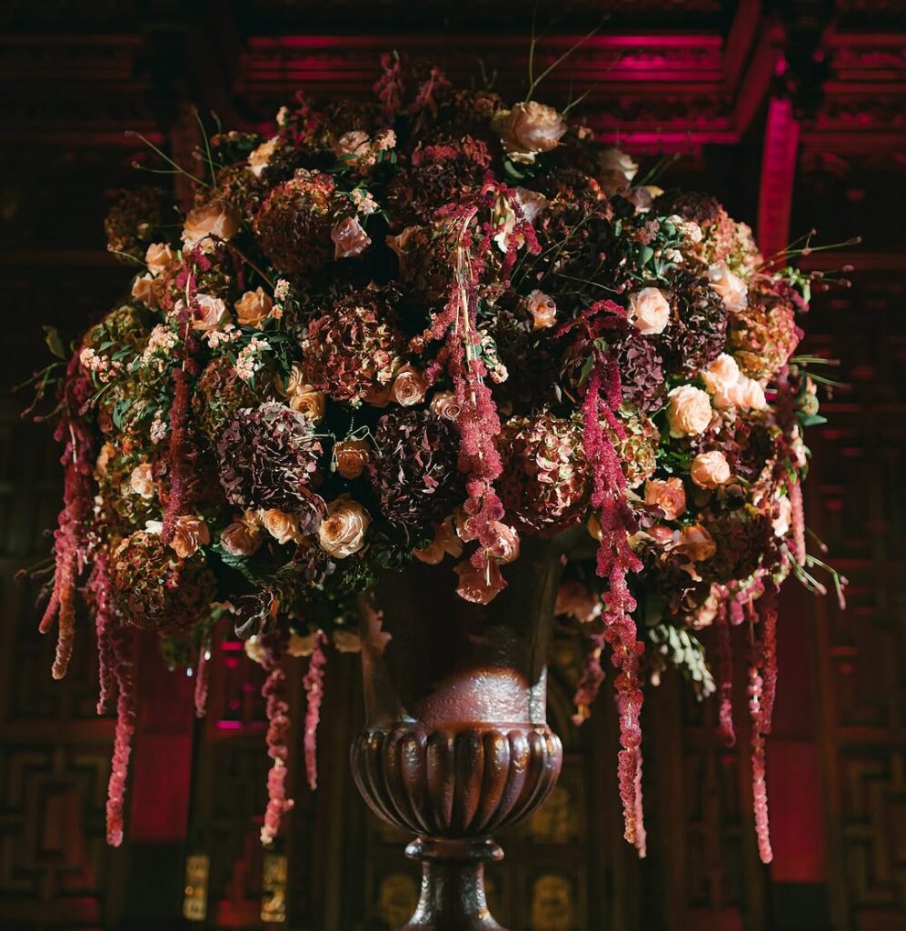 A grand floral arrangement in a large, ornate urn, showcasing vibrant hydrangeas, roses, and cascading amaranthus in rich autumnal colours.